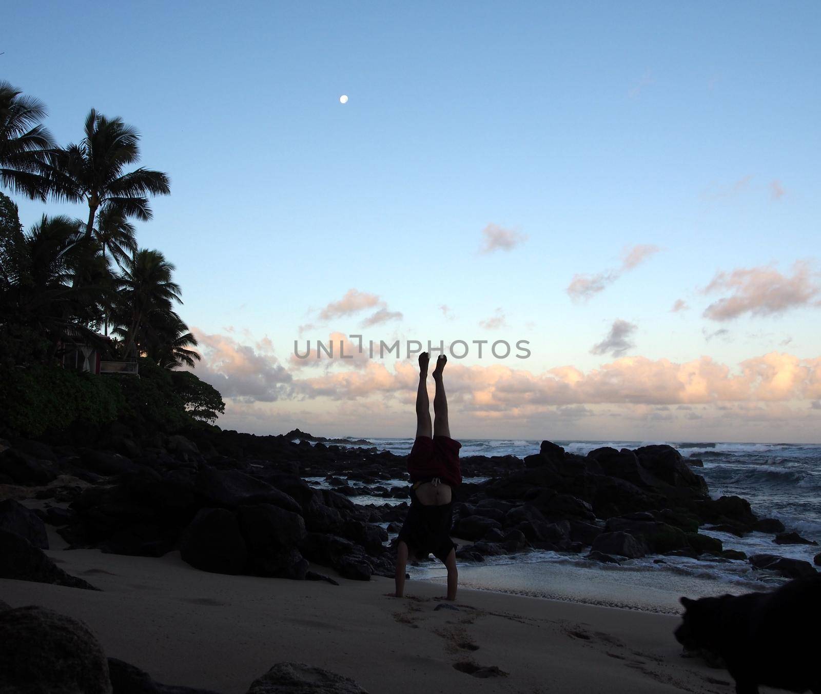 Man Handstanding on beach at dawn as wave crash on the North Shore and moon in the sky of Oahu, Hawaii.                    