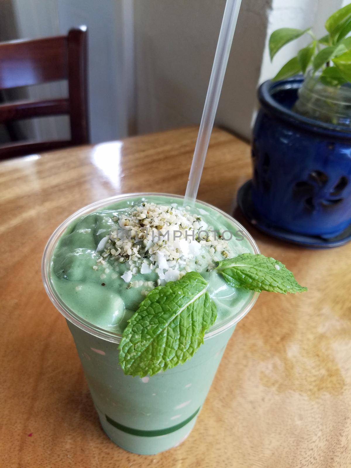 Veggin’ Out Smoothie in plastic cup with straw featuring Coconut water, bananas, avocado (when available), kale, spirulina, tahini, hemp seeds, flax meal and honey on a wood table.  (Smooth über healthy goodness!)