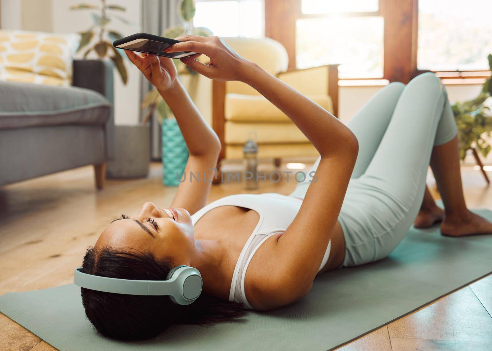 Downloading a few cool songs for her next session. a sporty young woman wearing headphones and using a cellphone while exercising at home. by YuriArcurs