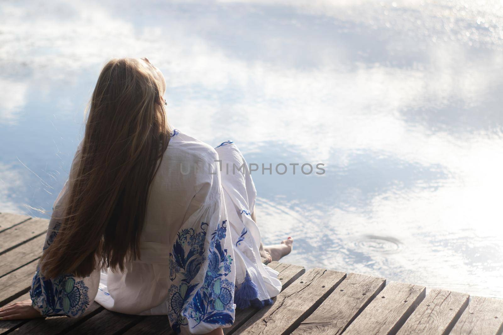 a girl in an embroidered Ukrainian shirt sits on the pier, the reflection of clouds in the water of the lake. On the shore of the sky. vyshyvanka day. freedom. patriot by oliavesna
