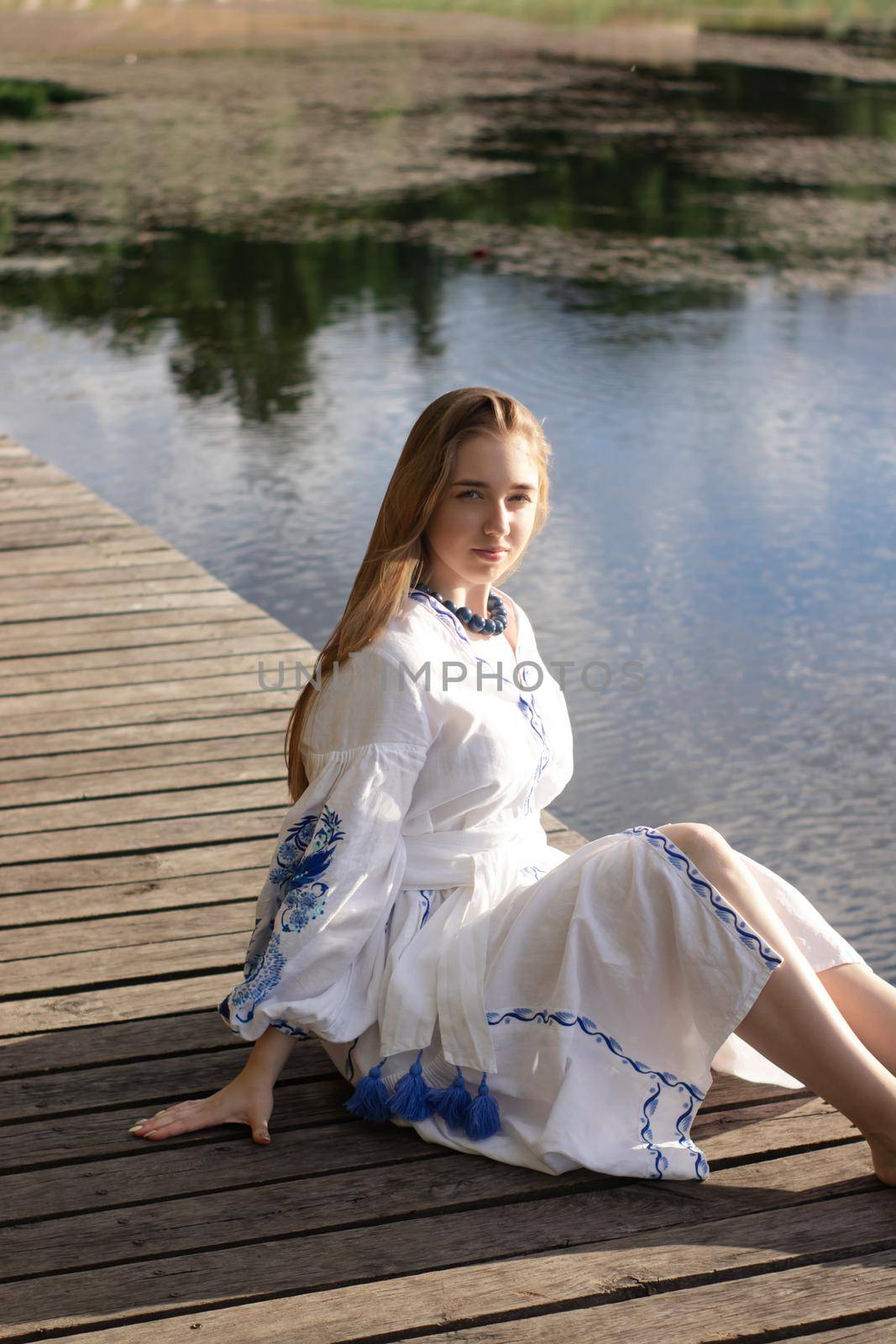 Girl in embroidered national Ukrainian costume on a pier on the shore of the lake. Independence day of ukraine, constitution, vyshyvanka day. young woman in blue dress outdoors by oliavesna