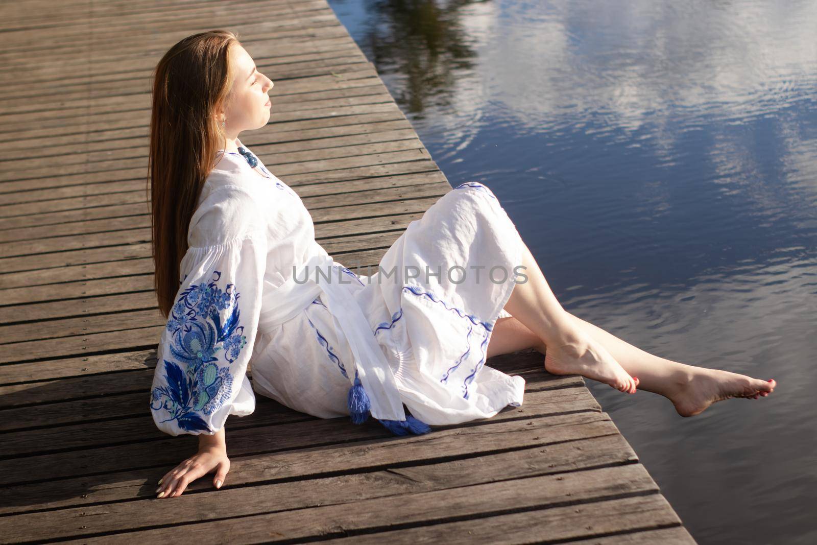 a girl in an embroidered Ukrainian shirt sits on the pier, the reflection of clouds in the water of the lake. On the shore of the sky. vyshyvanka day. freedom. patriot by oliavesna