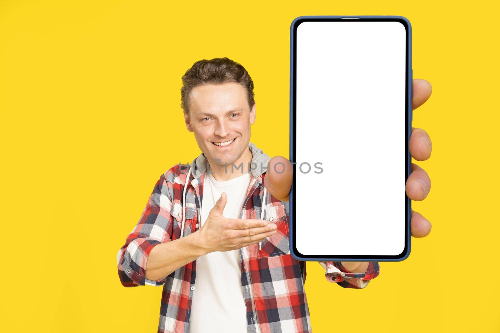 Introducing new app happy holding giant, huge smartphone with white screen blond man, wearing red plaid shirt. Man with phone display mock up isolated on yellow background. Mobile app advertisement.