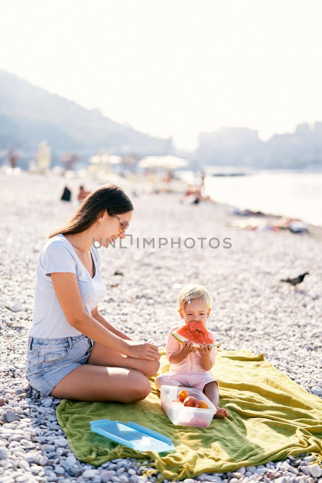 Smiling mom looking at baby girl eating watermelon on the beach. High quality photo