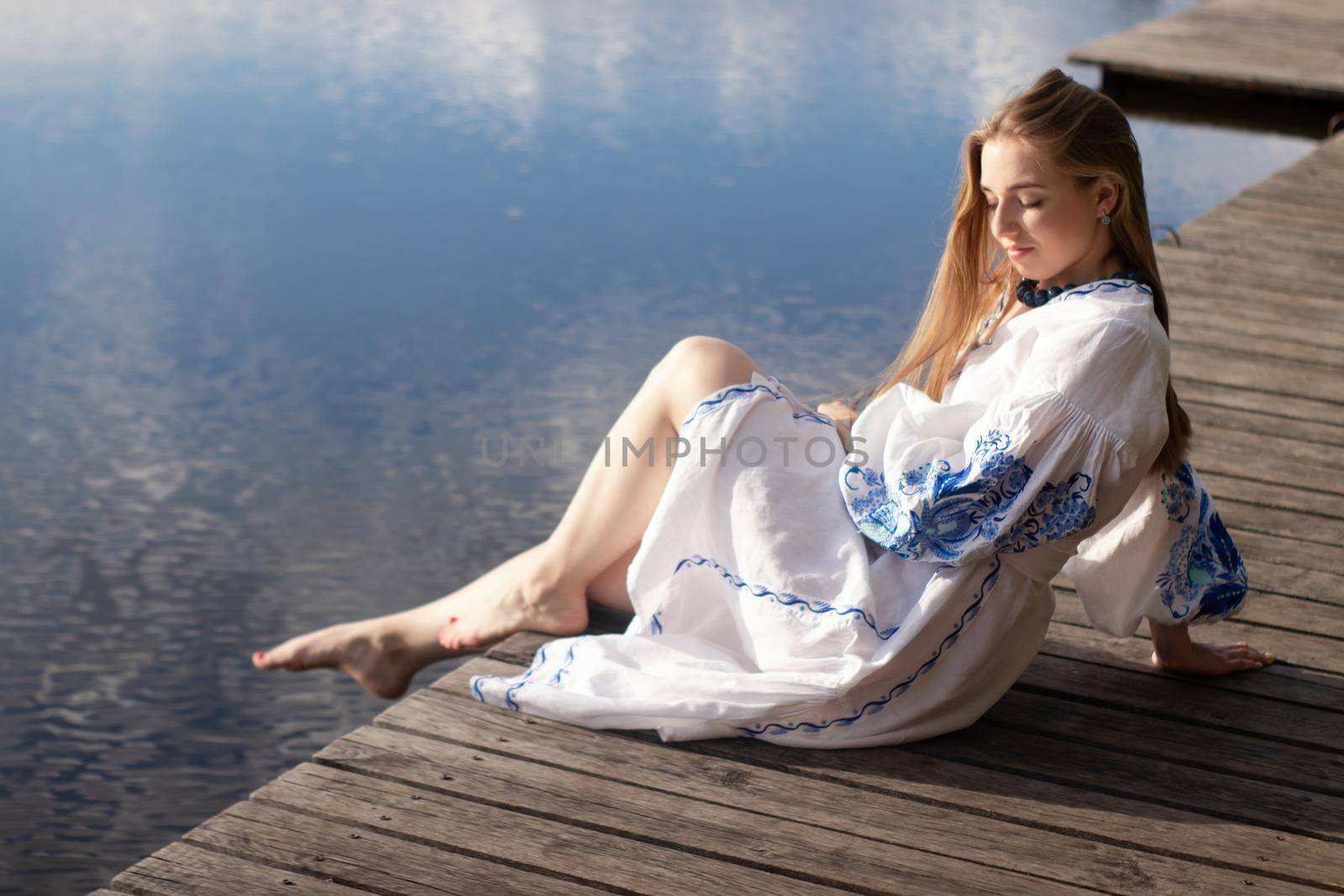 Girl in embroidered national Ukrainian costume on a pier on the shore of the lake. Independence day of ukraine, constitution, vyshyvanka day. young woman in blue dress outdoors by oliavesna