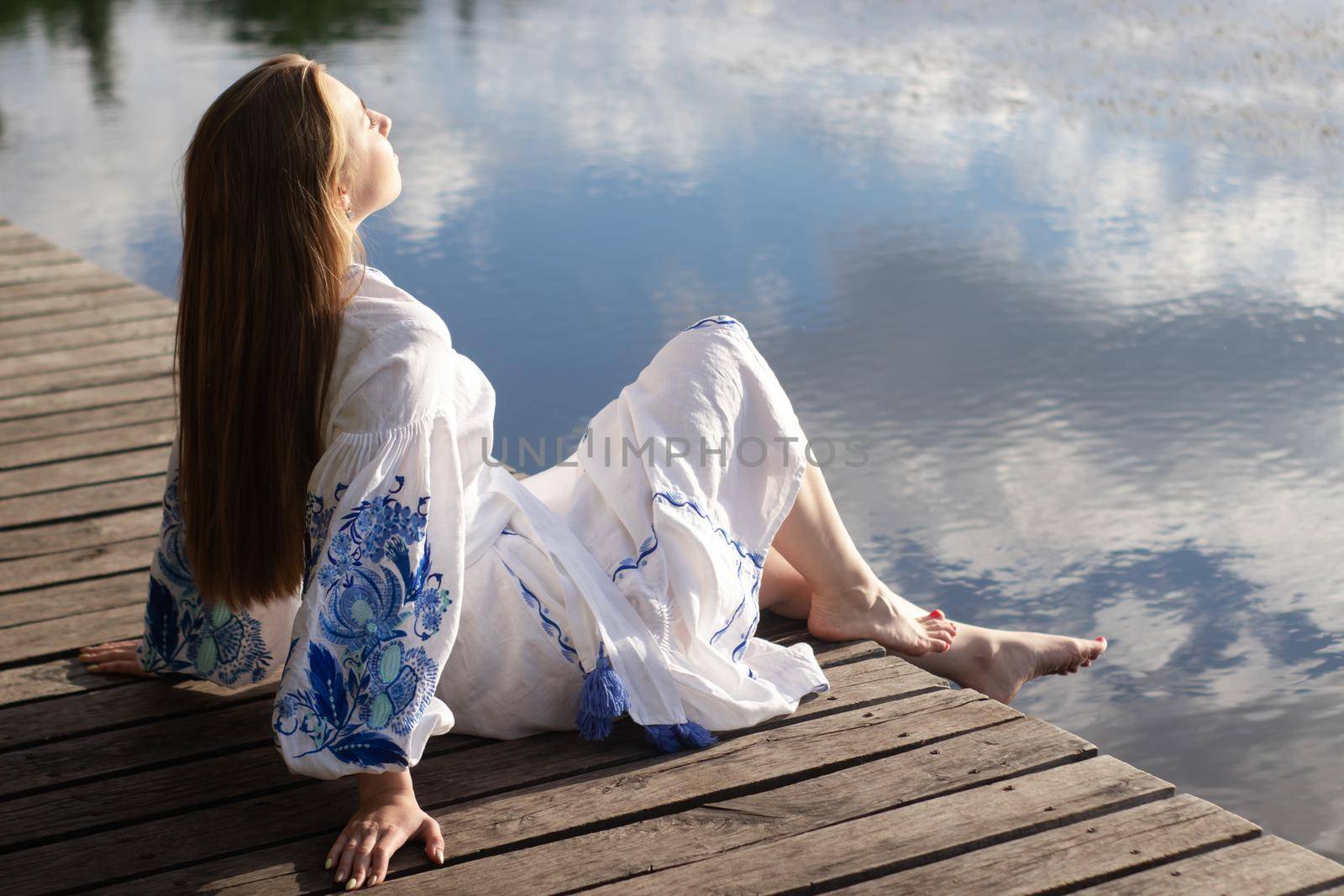 Girl in embroidered national Ukrainian costume on a pier on the shore of the lake. Independence day of ukraine, constitution, vyshyvanka day. young woman in blue dress outdoors by oliavesna