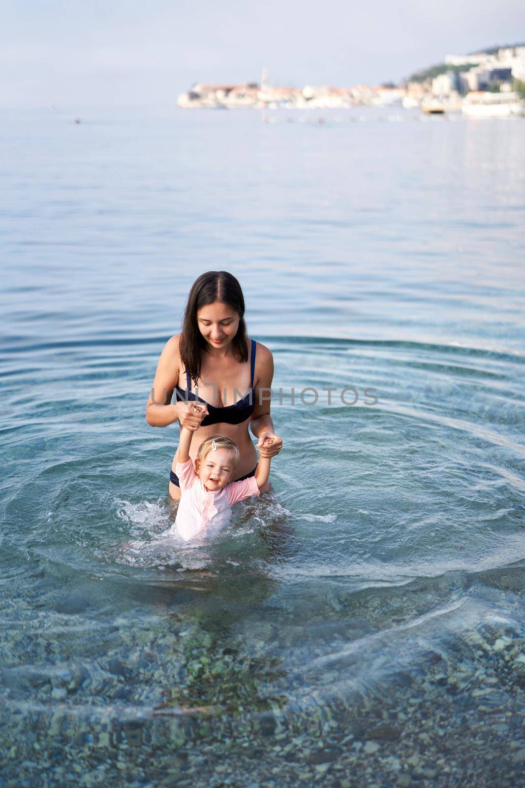 Smiling mother holding little girl hands in the sea on a shallow by Nadtochiy