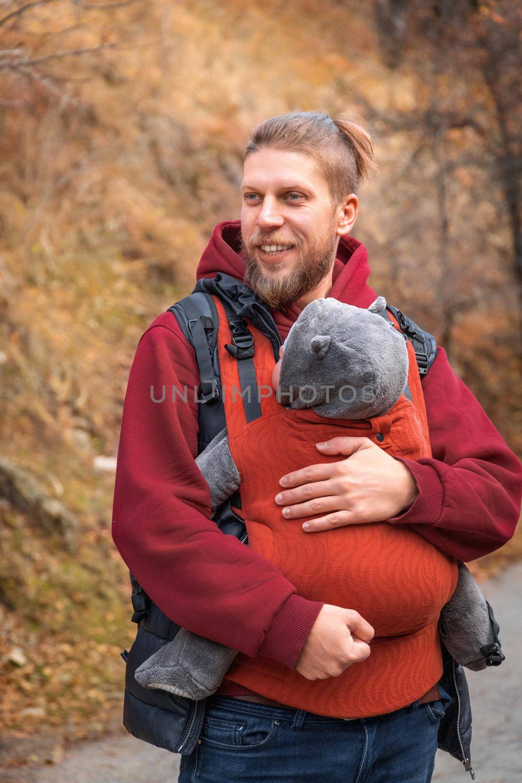 Adorable smiling bearded babywearing father with his baby in sling autumn walking.
