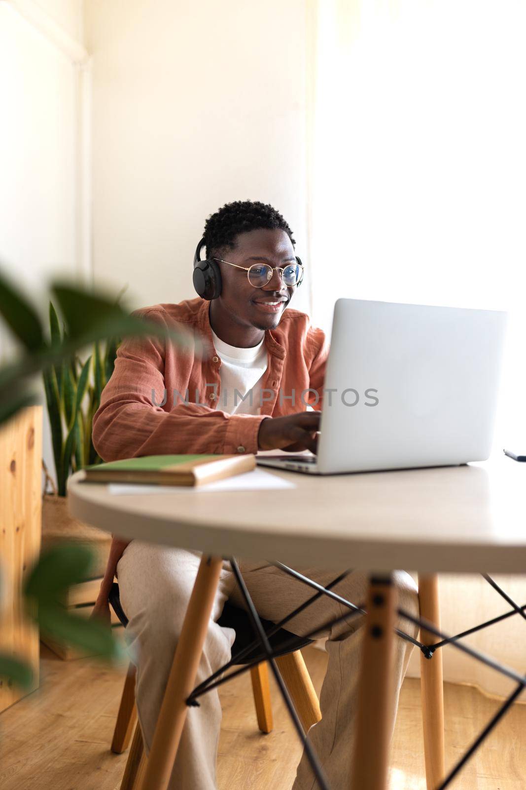 Vertical portrait of young African American man wearing headphones working, studying at home using laptop. Copy space. by Hoverstock