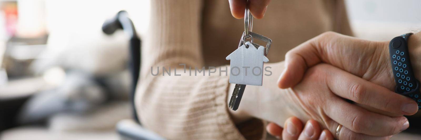 Close-up of realtor give keys from new accommodation to woman in wheelchair, kind handshake in agreement. Disability, government help, real estate concept