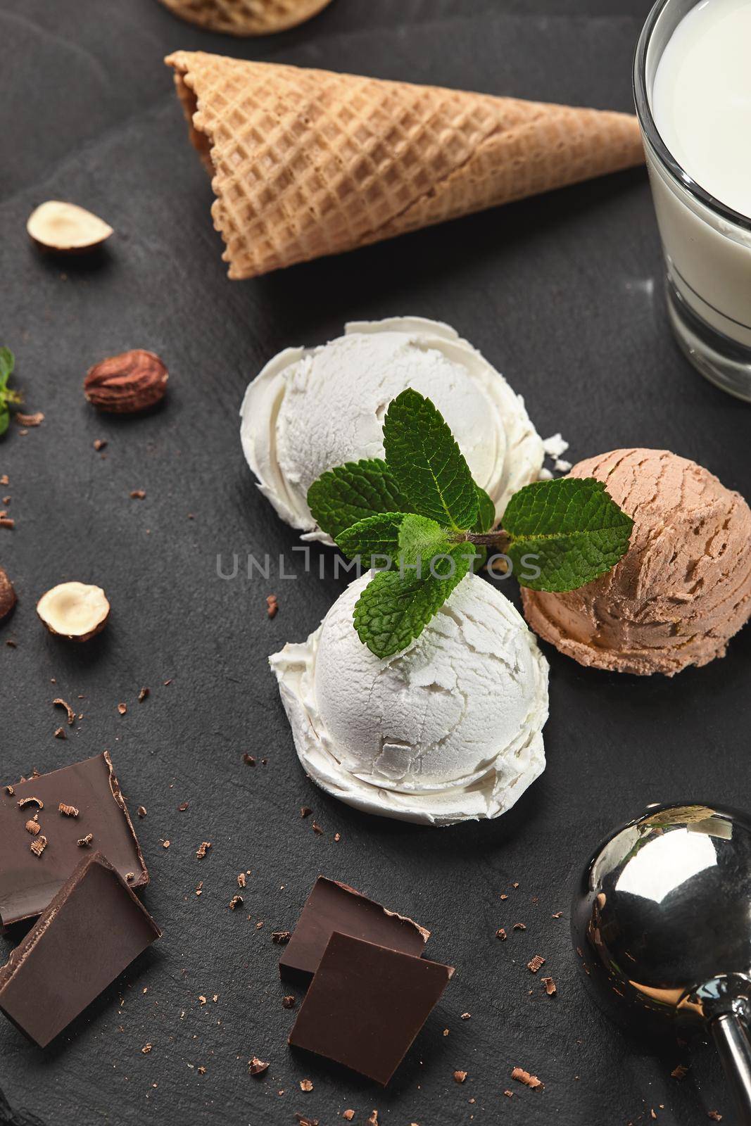 Top view of exquisite creamy and chocolate ice cream decorated with mint, a glass of milk, metal scoop, waffle cone with dark chocolate and hazelnuts nearby, served on a stone slate over a black background. Close-up.