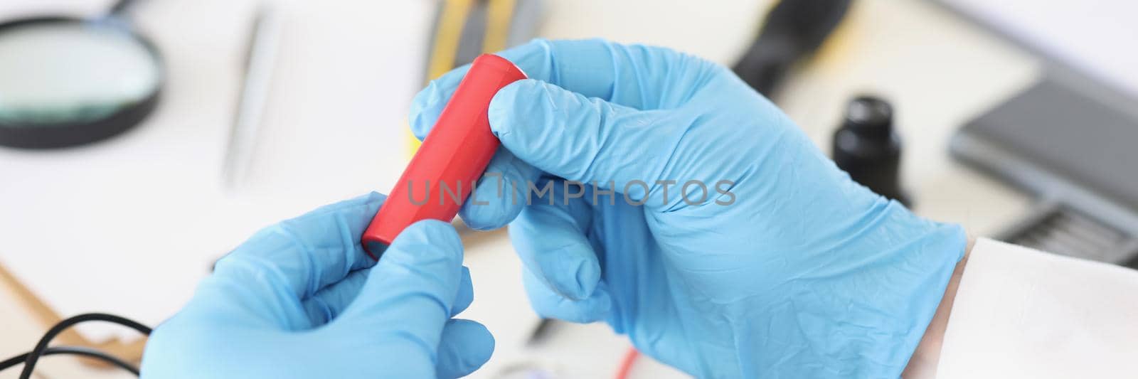 Close-up of male in blue gloves holding red battery piece, repairing broken or damaged device by himself. Workplace full with tools. Handyman, fix concept