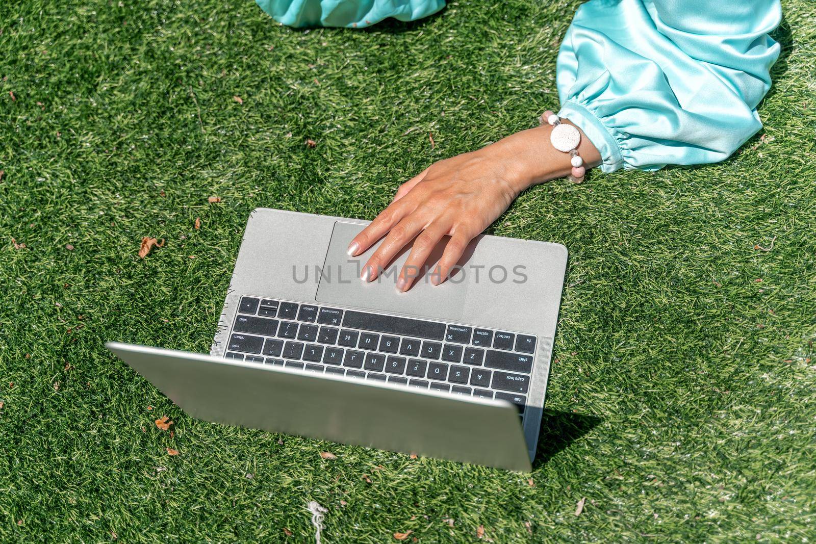 a young beautiful woman with blond curly hair in glasses and a blue dress sits on the grass in nature and uses a laptop