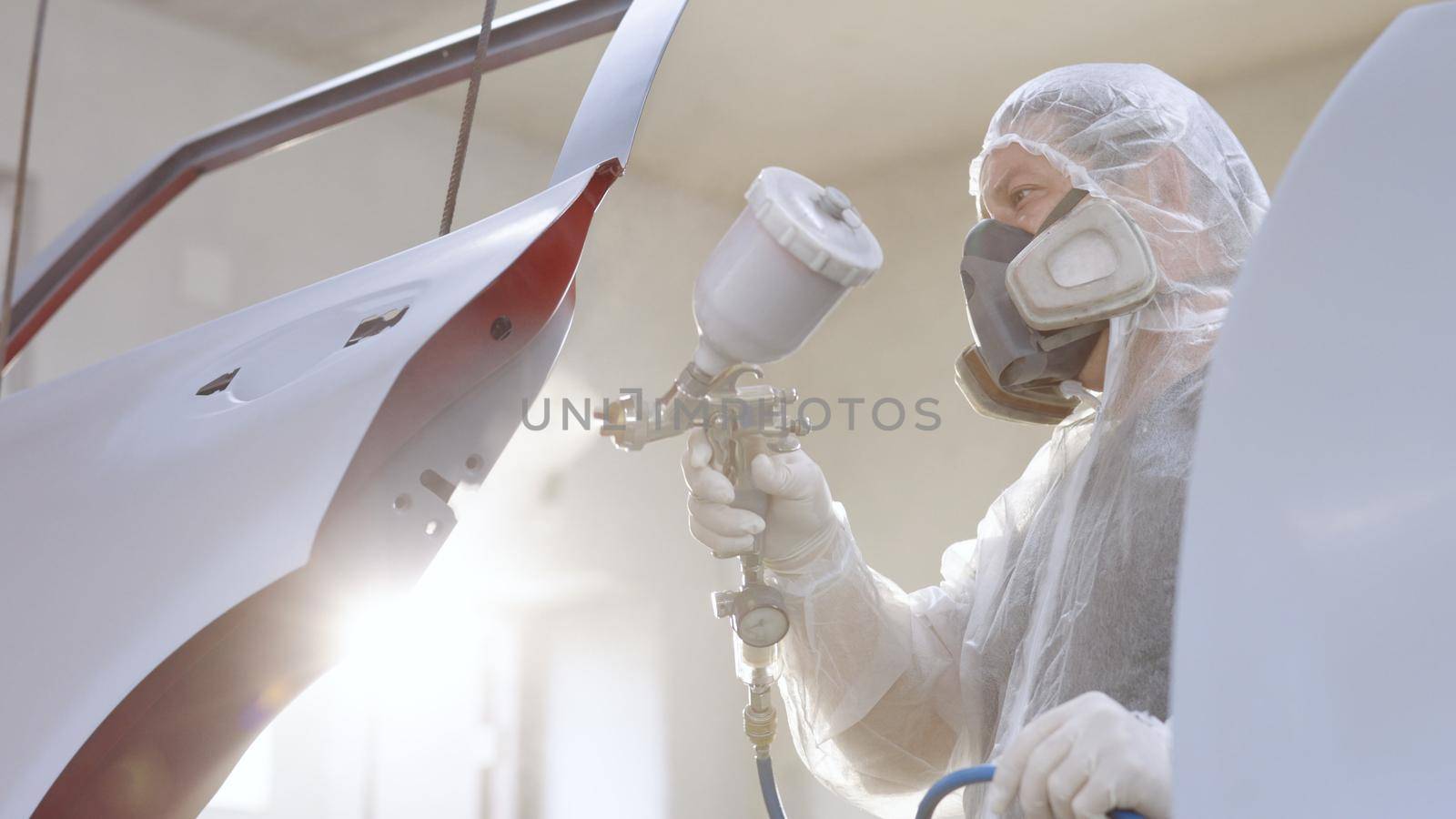 Master is painting part of automobile in car service. He is using spraying machine, holding it in hand, dressed in protective uniform. Modern car painting. by uflypro