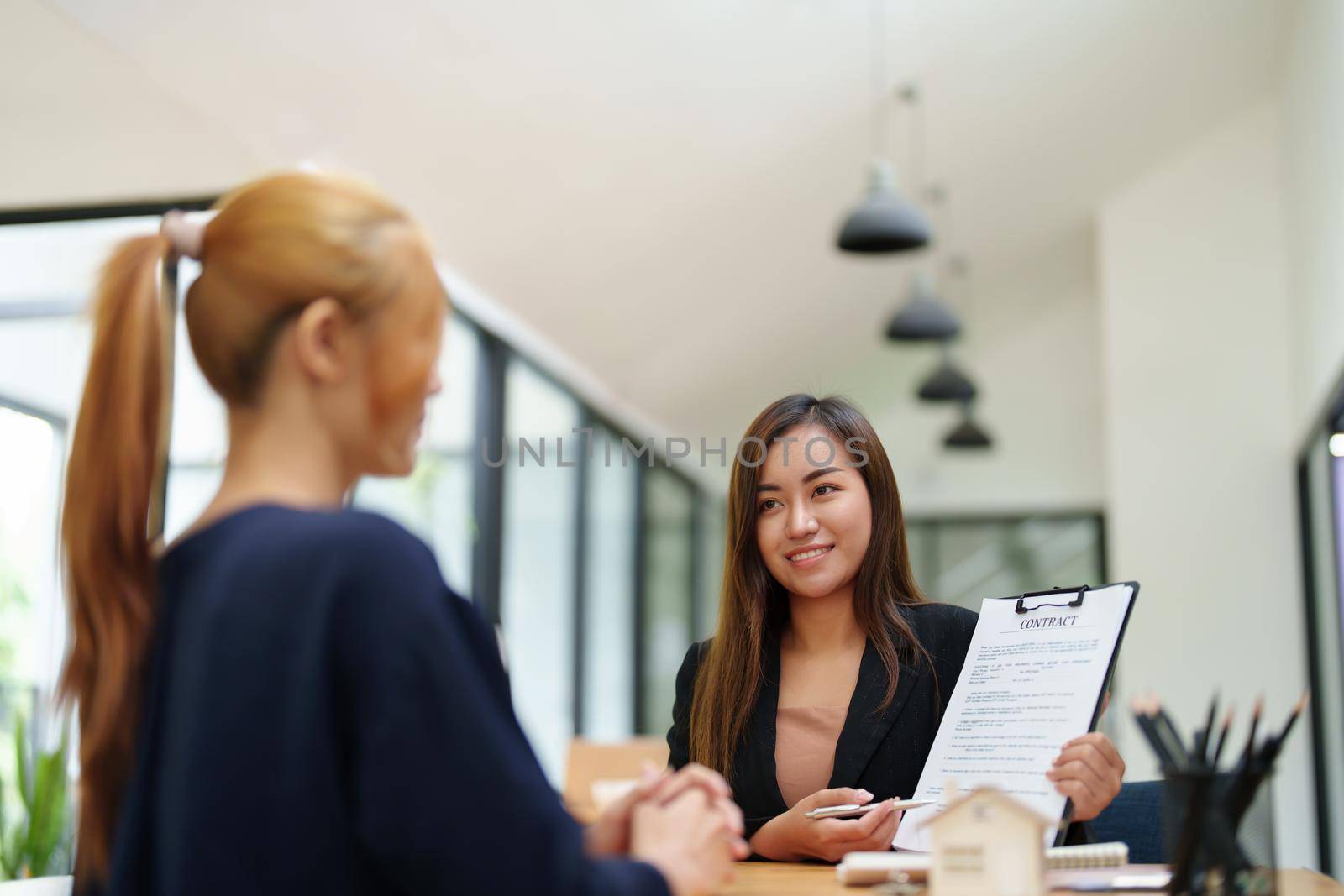Portrait of an Asian female bank employee asking a customer to read the contract before signing to agree to buy a house, real estate concepts by Manastrong