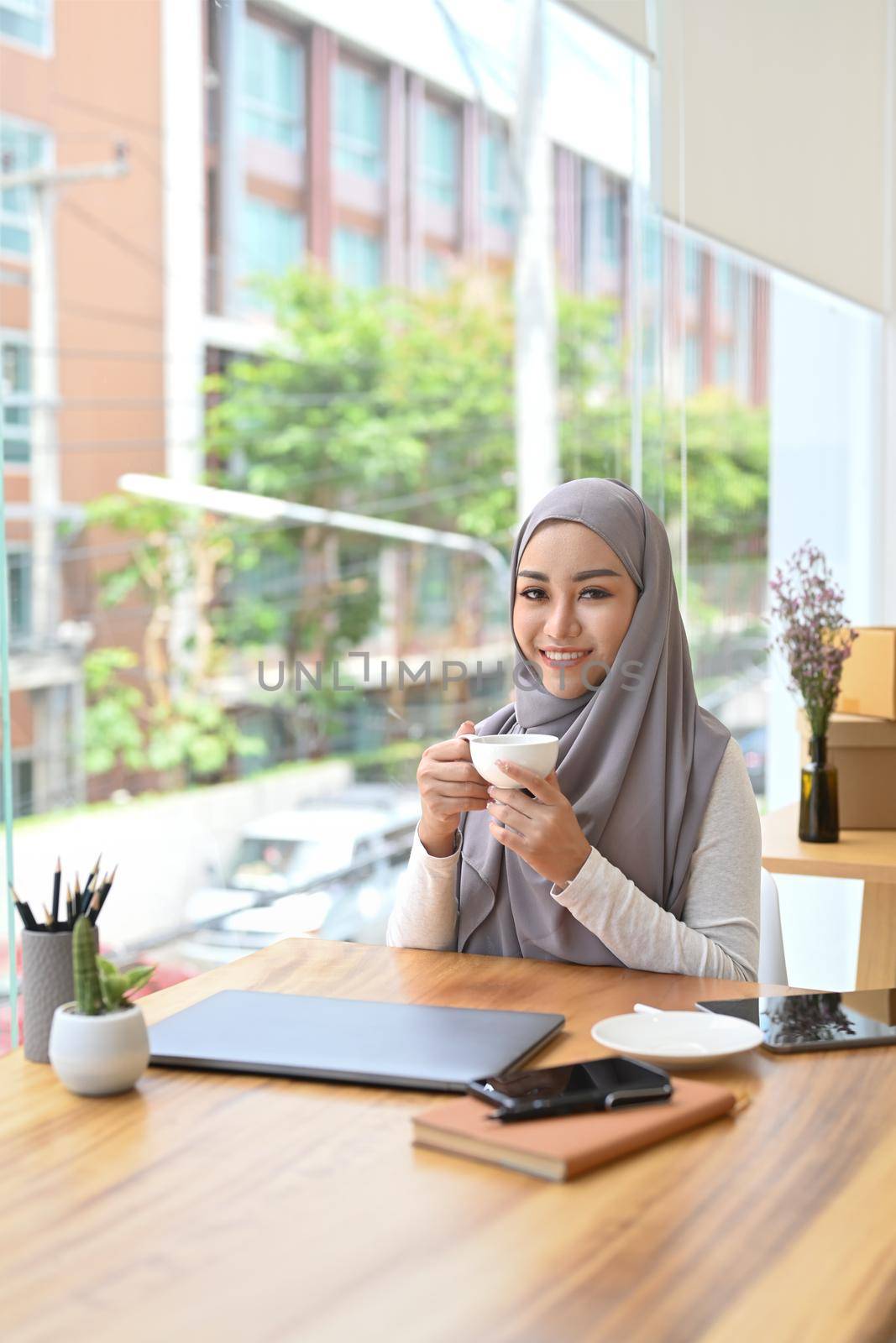 Smiling muslim businesswoman in hijab sitting in her office and enjoying morning coffee.
