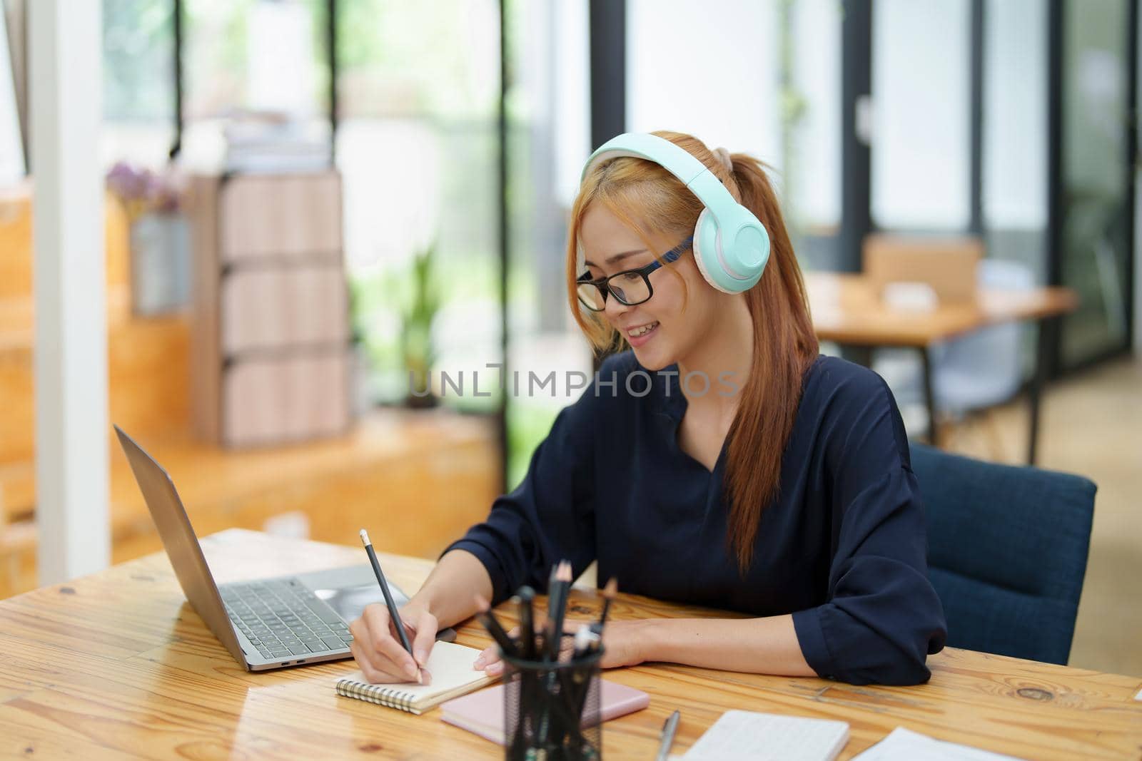 Portrait of a young Asian woman sitting at work wearing headphones over her ears to listen to music for pleasure by Manastrong