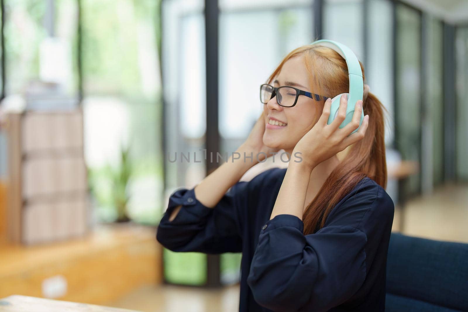 A portrait of a young Asian woman with blonde hair wearing over-ear headphones listening to music to relax while taking a break from boring day activities by Manastrong