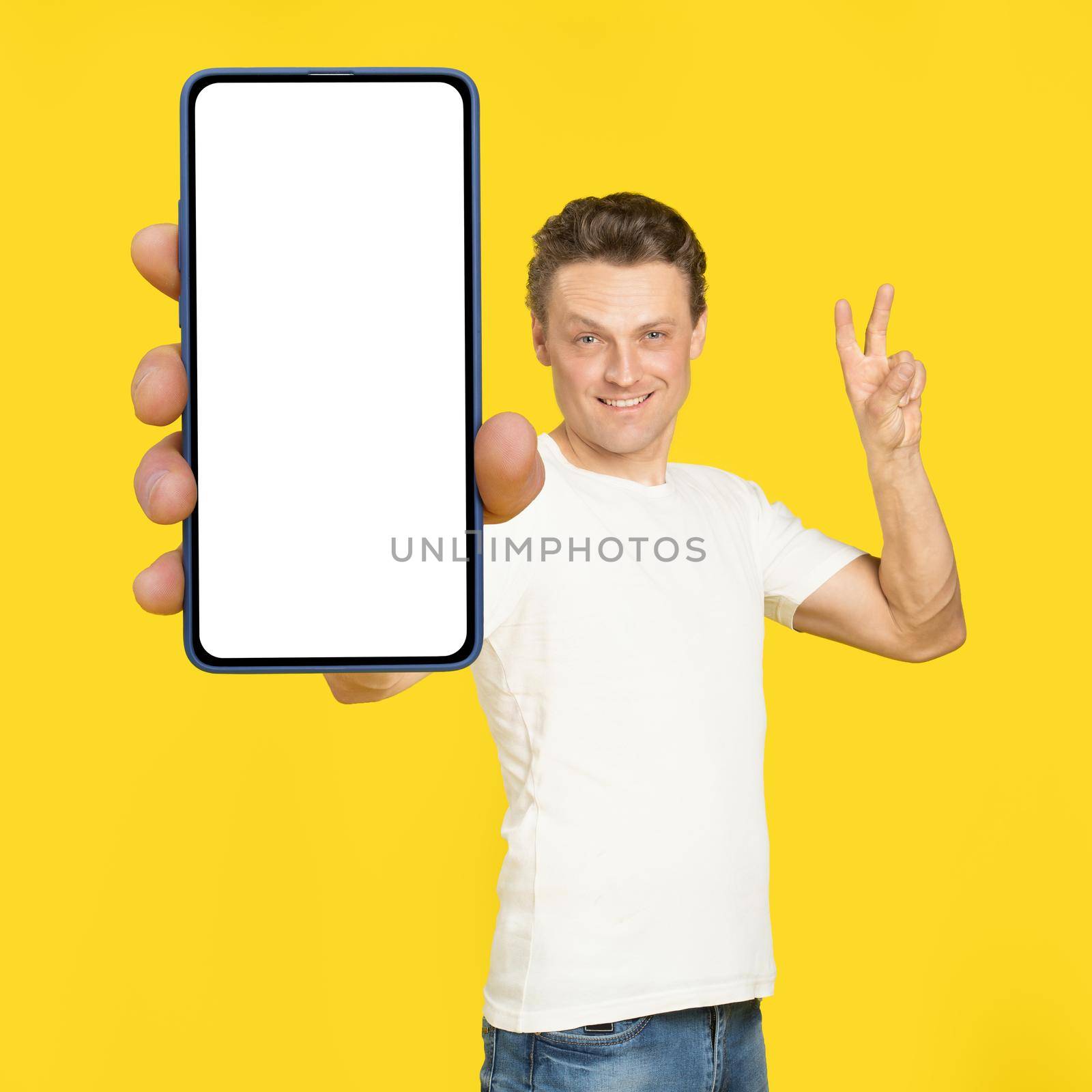 Gesturing V or victory handsome man holding huge smartphone with white screen wearing white t-shirt and jeans isolated on yellow background. Mobile app advertisement, great offer.