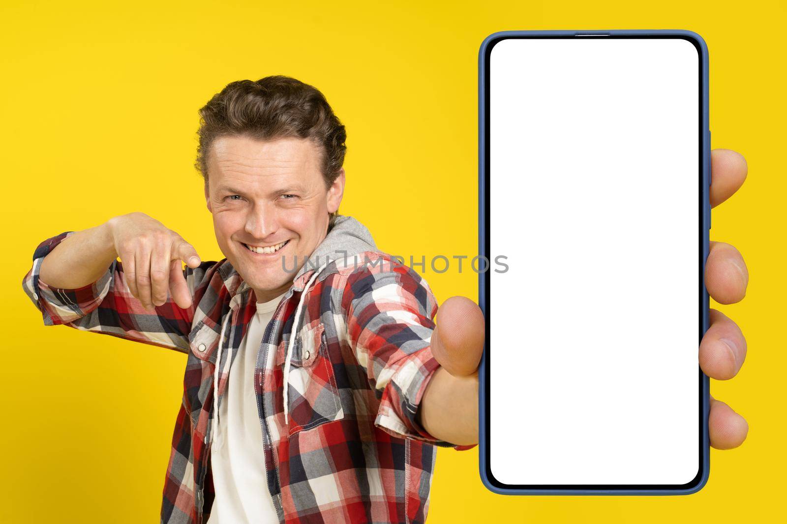 Pointing at big smartphone in hand with white screen blond caucasian man, wearing red plaid shirt. Man with phone display mock up isolated on yellow background. Mobile app advertisement.