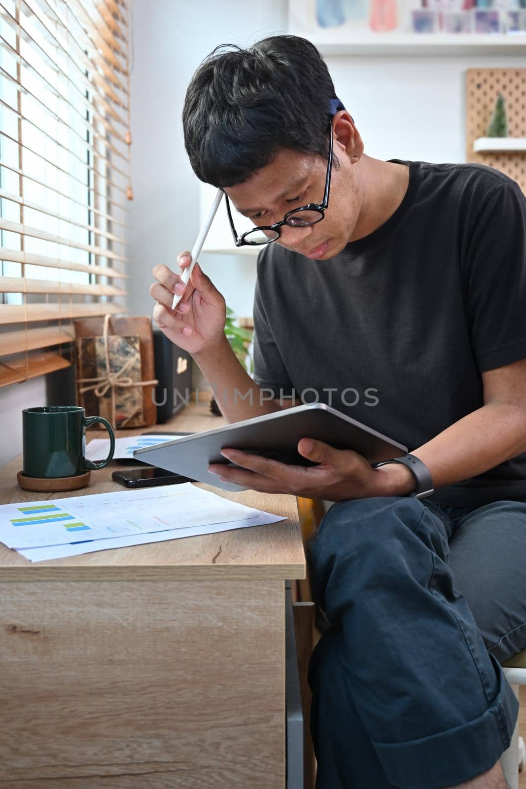 Young man sitting in home office and analyzing financial data on digital tablet. by prathanchorruangsak