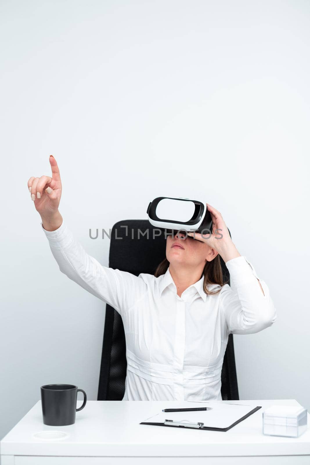 Businesswoman Wearing Virtual Reality Goggles And Learning Professional Skill Through Futuristic Simulation. Woman Gesturing While Sitting At Desk During Modern Training. by nialowwa