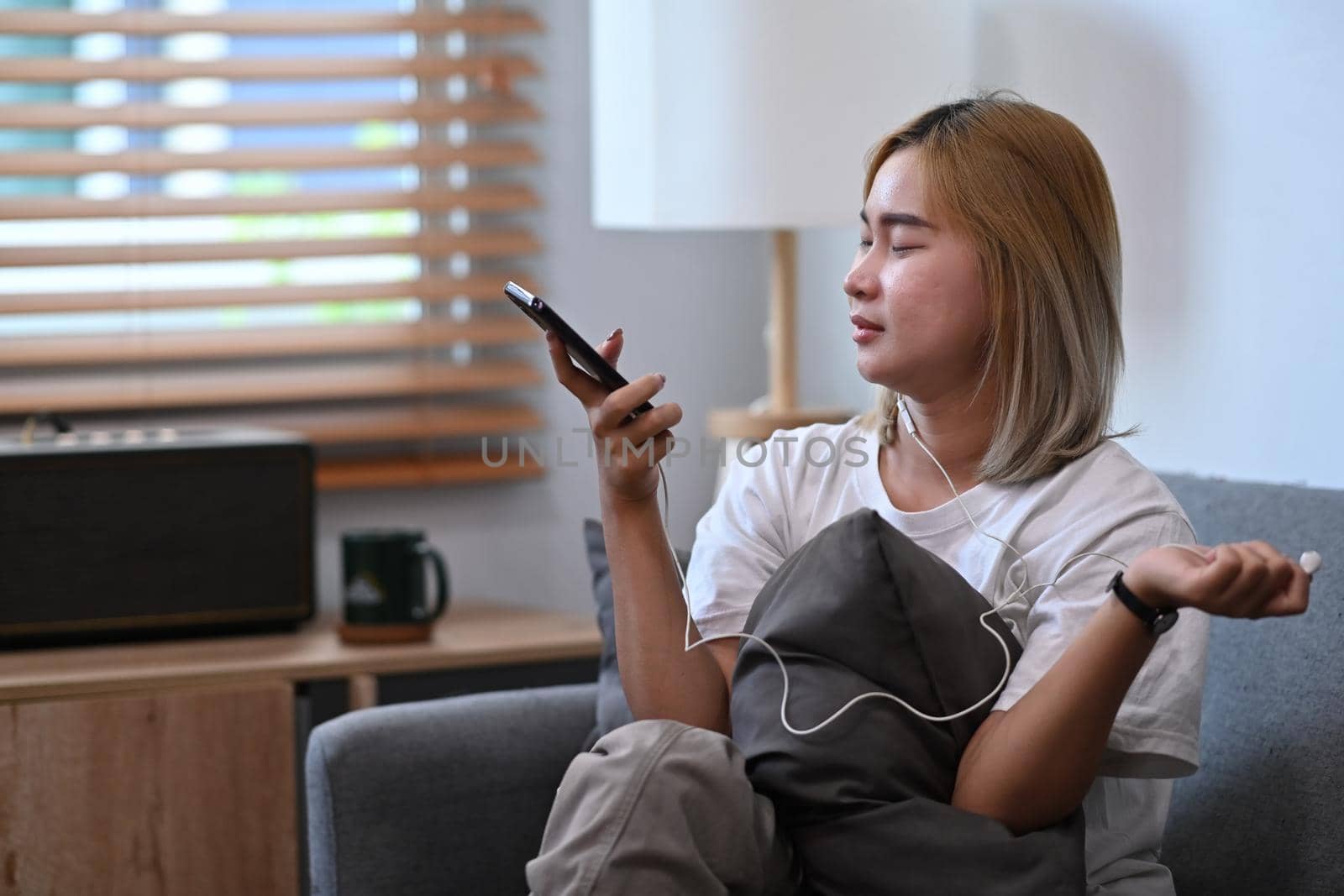 Happy asian woman wearing earphone and using smart phone on couch.