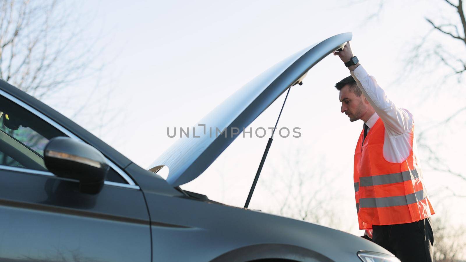 Disappointed man in formal outfit opening bonnet of broke down car to check engine. Bearded businessman is getting late to working meeting because of troubles on road.