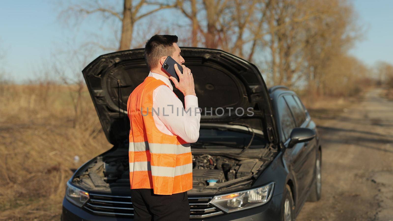 Stranded man with broken down car calls for help. Young man standing speak on phone calling car assistance services, near the broken car opened the hood look road help repair. Emergency stop sign by uflypro