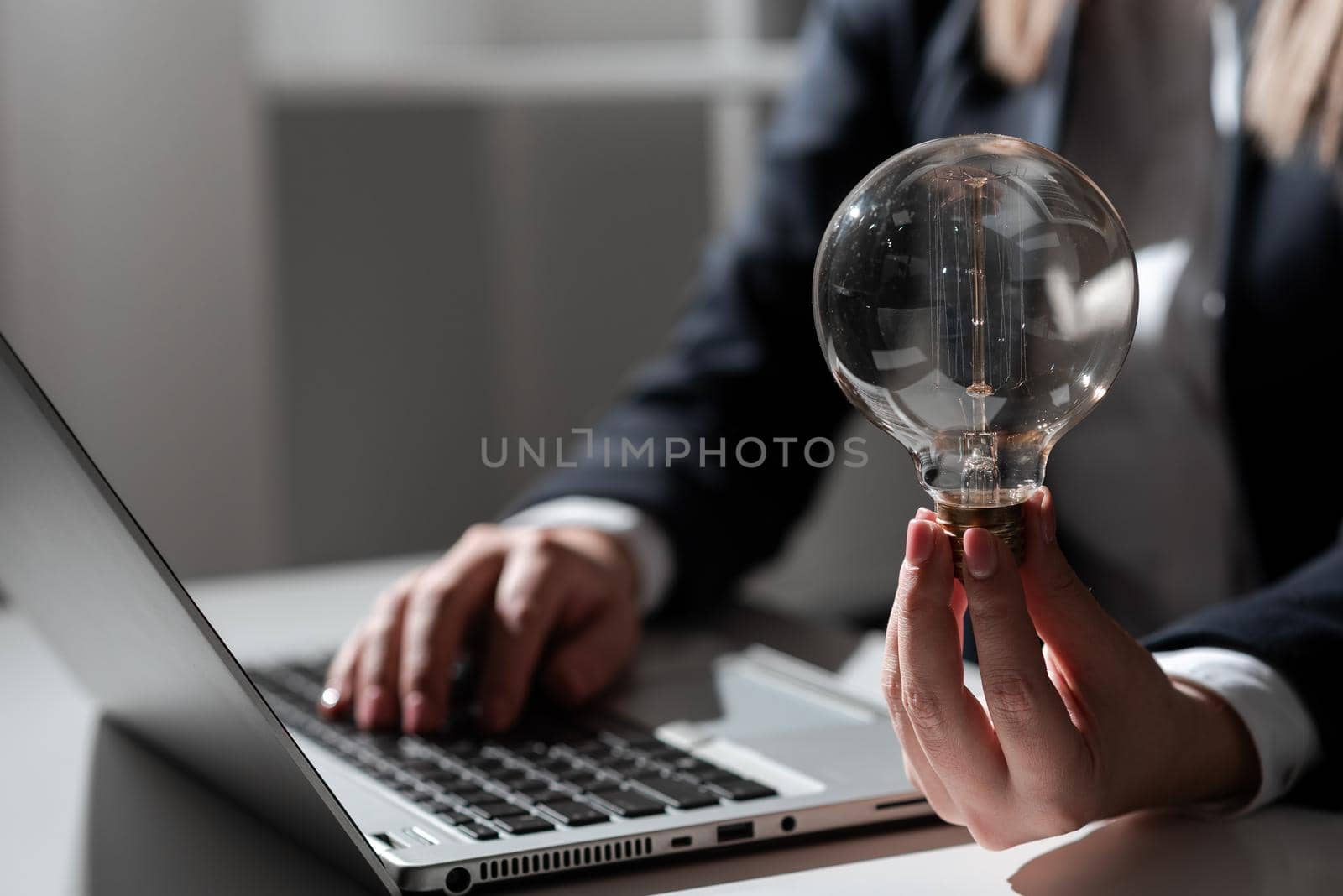 Businesswoman Typing Recent Updates On Lap Top On Desk Holding Lightbulb. Woman In Office Writing Important Message On Computer. Executive Inserting Crutial Data Into Pc. by nialowwa