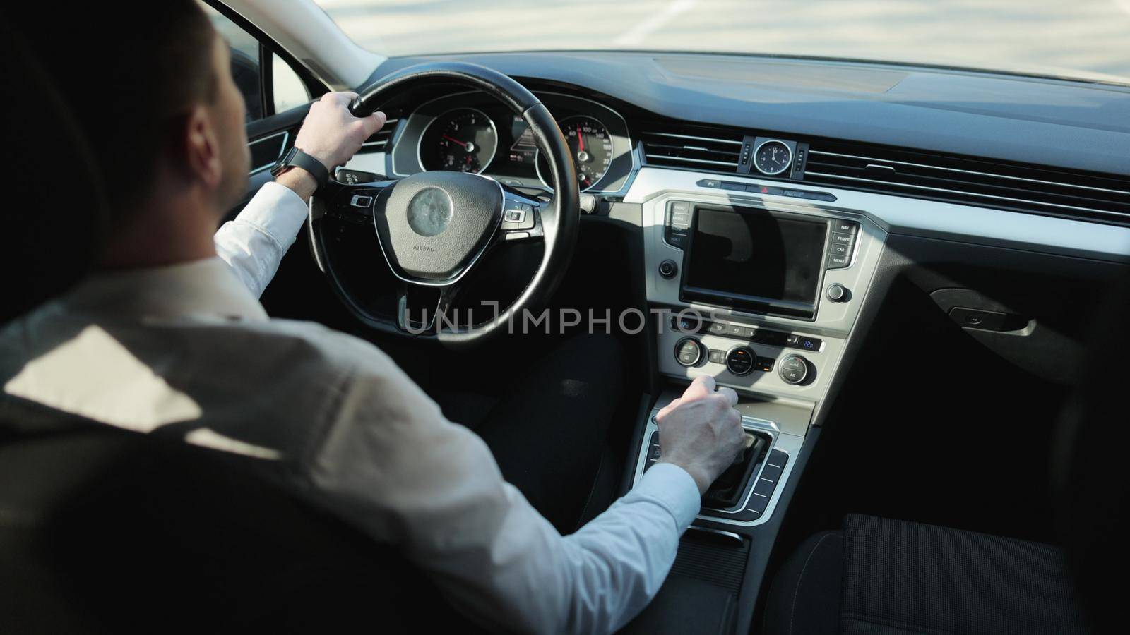 Caucasian bearded adult man in white shirt driving luxury car in sunny day across the highway. Side back view. Successful business people traveling lifestyle. Navigation. Highway transportation.