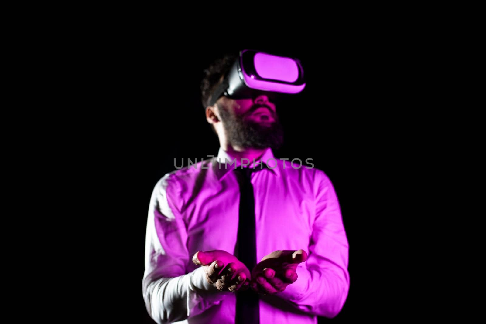 Bearded Businessman Wearing Virtual Reality Goggles And Enjoying Simulator. Light Falling On Man Using Futuristic Gadget Gesturing And Presenting Modern Innovative Technology. by nialowwa