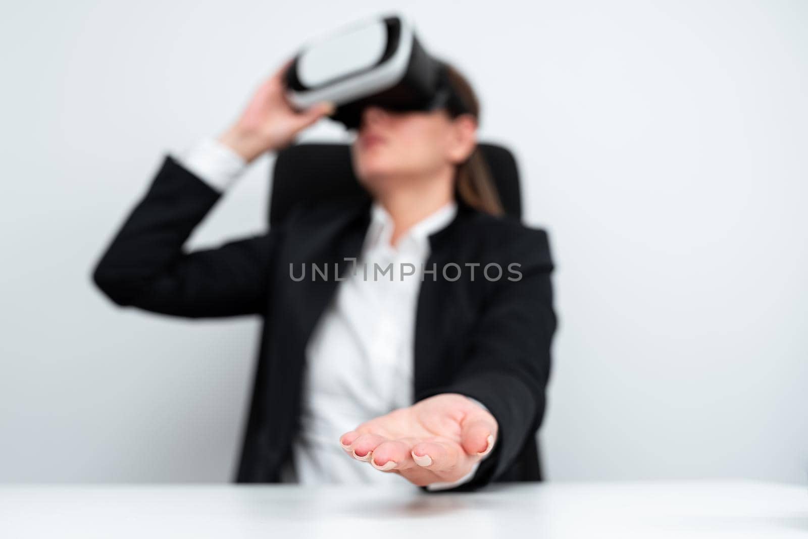 Female Executive Wearing Virtual Reality Simulator Gesturing During Professional Training. Businesswoman Wearing Suit Sitting At Desk And Presenting Futuristic Technology. by nialowwa