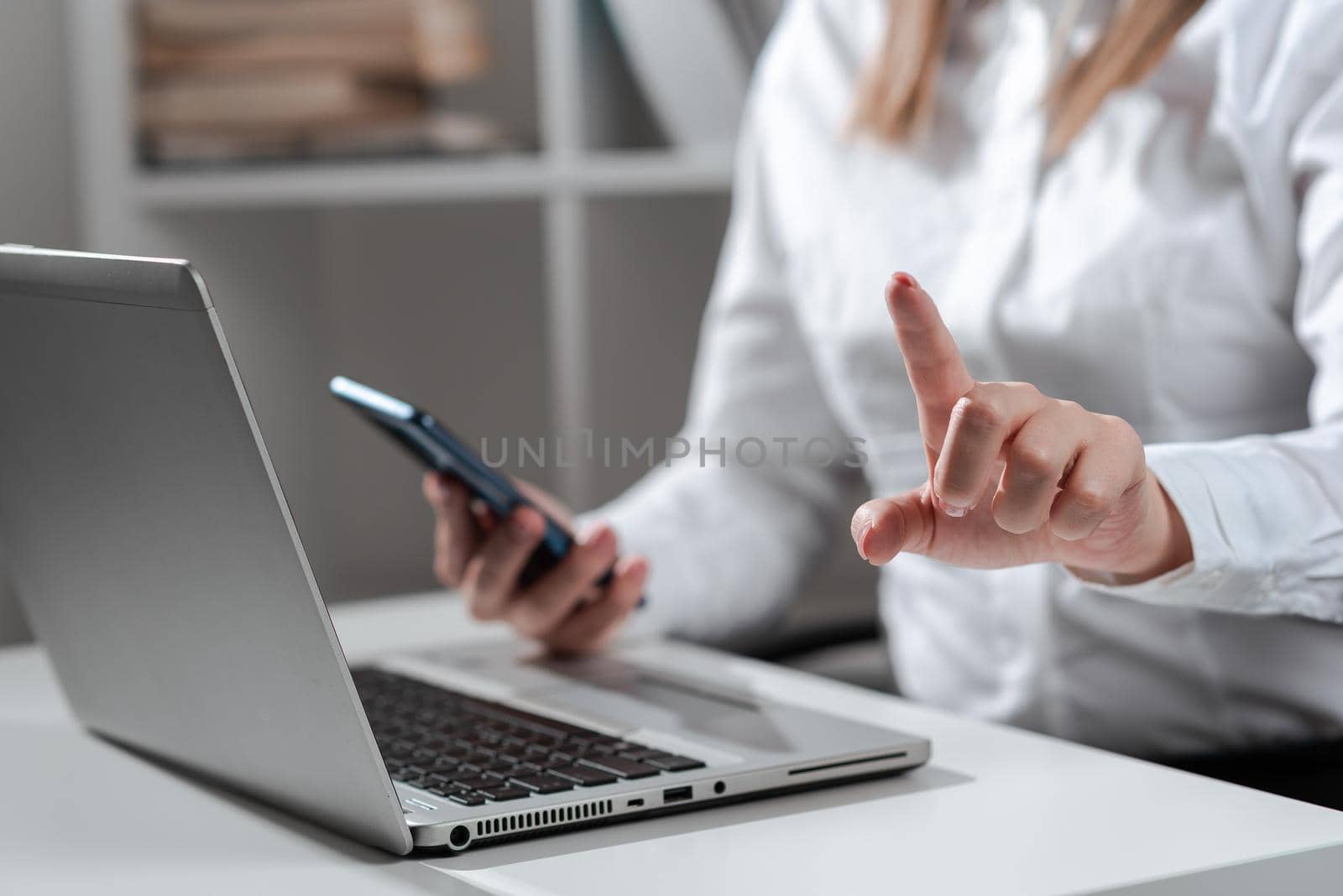 Businesswoman Holding Phone And Pointing With One Finger On Important Message On Desk With Lap Top. Executive In Suit Presenting Crutial Informations. by nialowwa