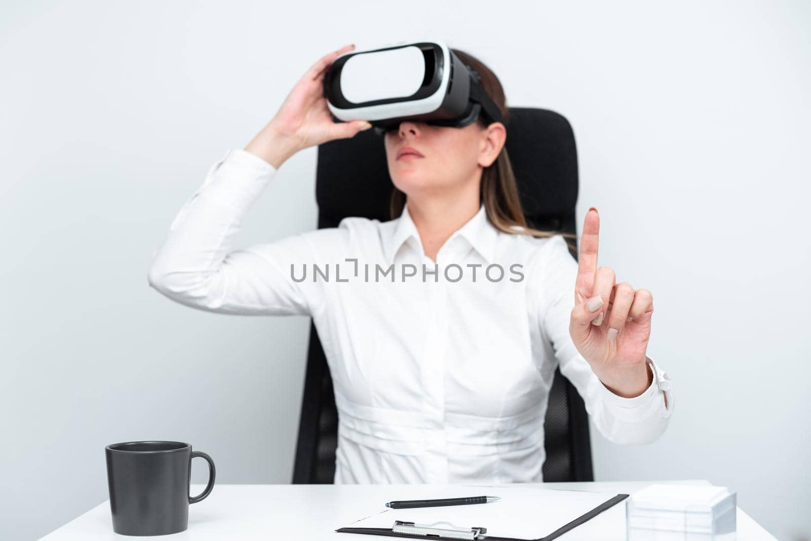 Businesswoman Wearing Virtual Reality Headset And Gesturing While Learning Professional Skill Through Simulation. Woman Sitting At Desk And Experiencing Modern Technology. by nialowwa