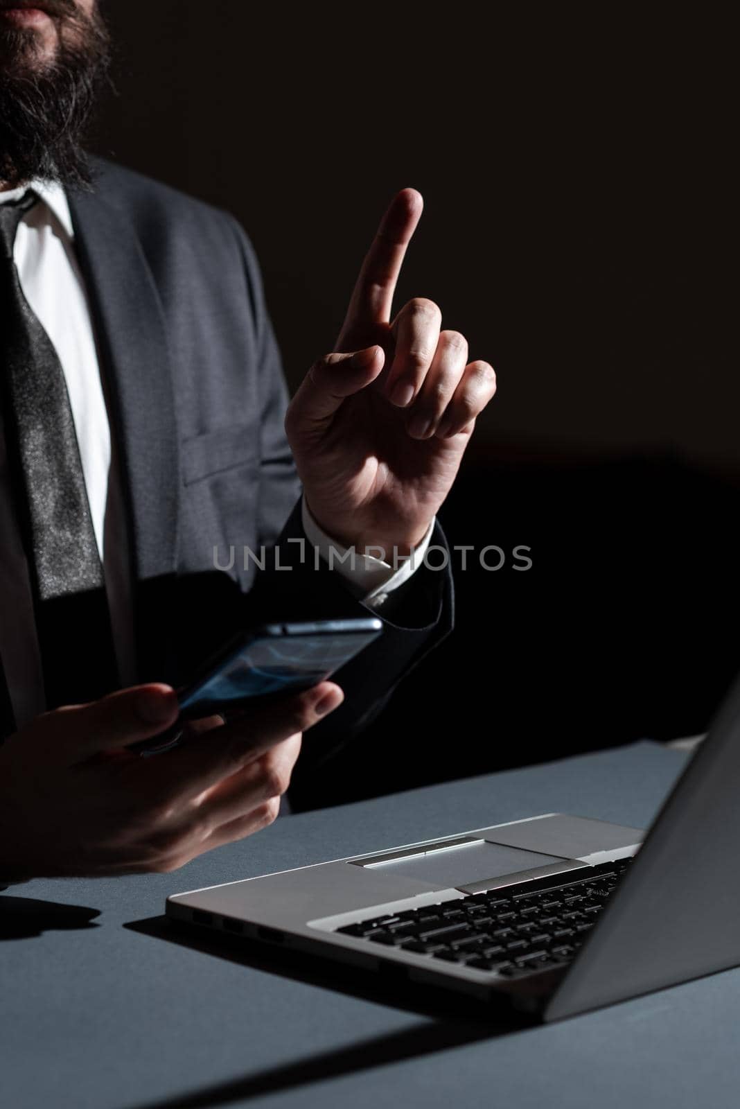 Businessman Holding Mobile Phone In Hand And Pointing With One Finger On Important Message On Desk With Lap Top. Man Having Cellphone And Presenting New Ideas With One Hand. by nialowwa