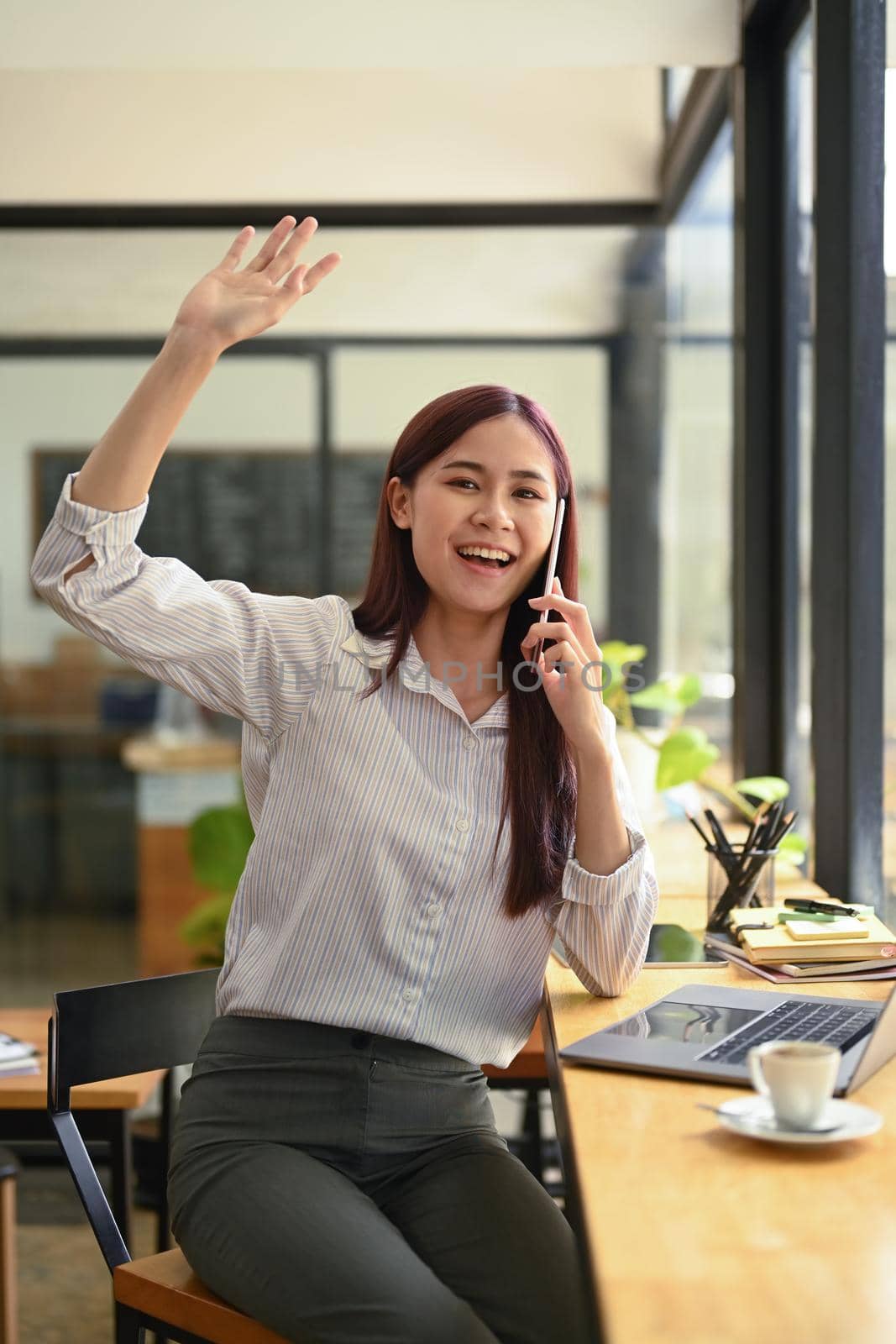 Friendly asian female employee talking on mobile phone and waving hands to greet her colleagues.