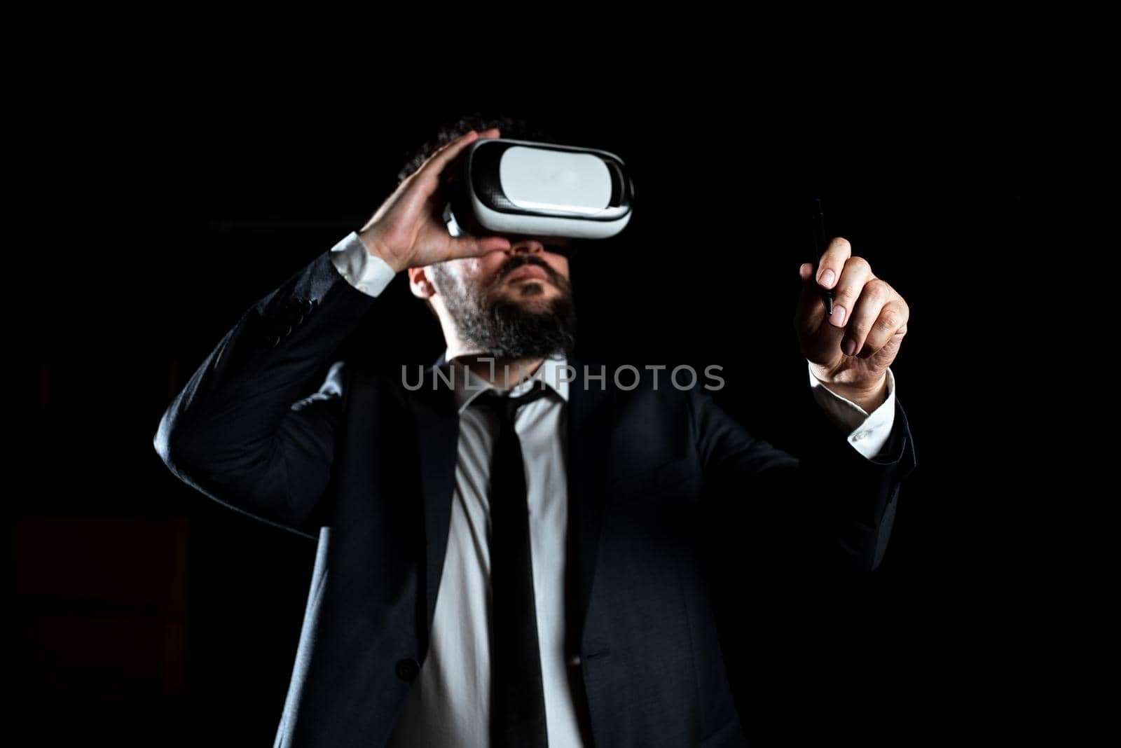 Male Professional Wearing Virtual Reality Goggles Holding Pen And Presenting Modern Technology Of Learning. Businessman Wearing Suit Taking Training Through Simulator. by nialowwa