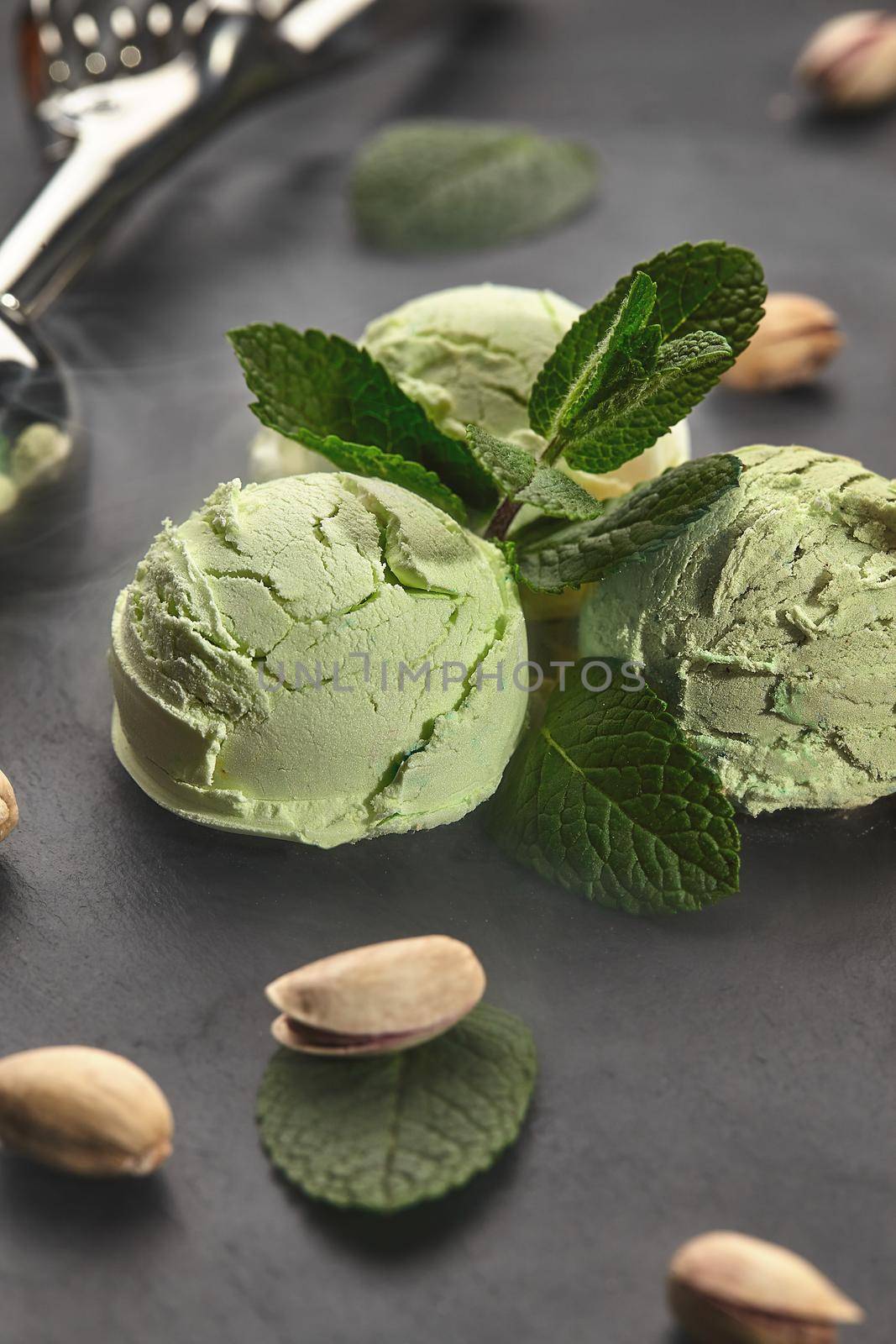 Close-up shot of a tasty pistachio ice cream decorated with mint, scattered pistachios are nearby, served with a metal scoop on a slate over a black background.