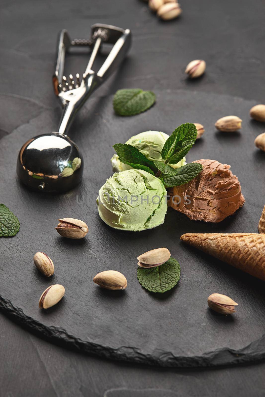 Delicious chocolate and pistachio ice cream decorated with mint, classic waffle cones with scattered nuts are nearby, served with a metal scoop on a stone slate over a black background. Close-up.