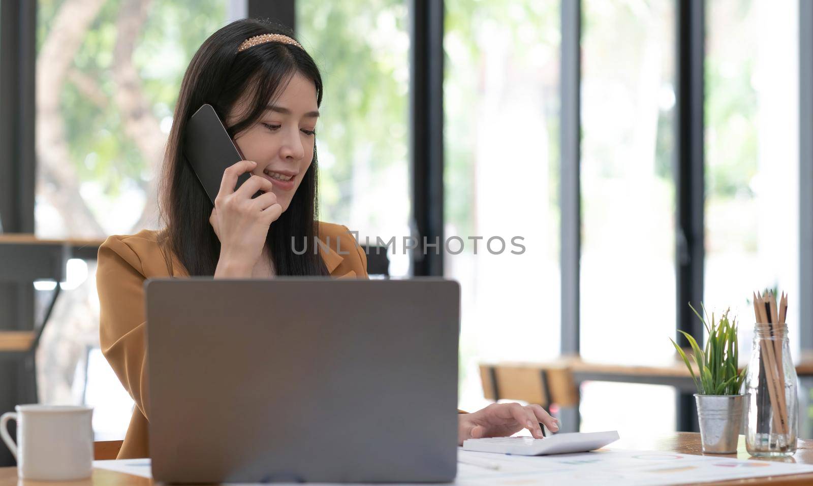 Beautiful Asian businesswoman analyzes charts using laptop calculator and talking the phone at the office. by wichayada