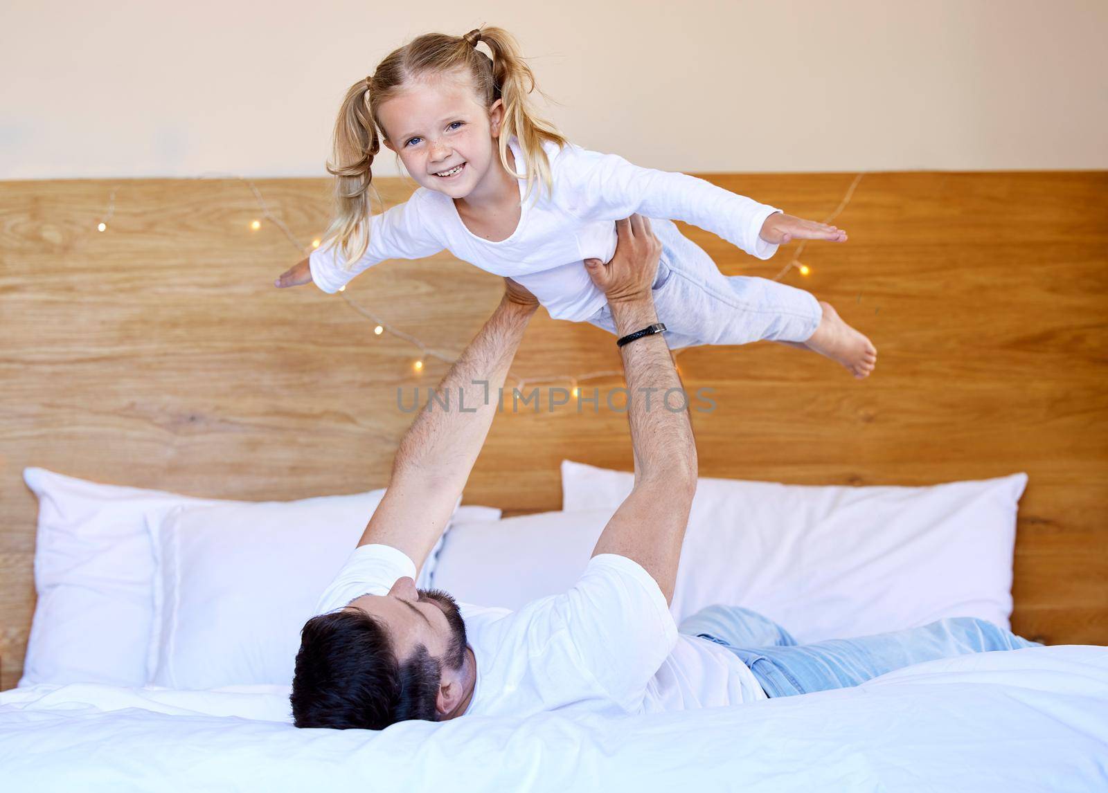 Adorable little girl bonding with her single father at home and pretending she can fly. Caucasian single parent holding and lifting his daughter in the air. Smiling child playing in the bedroom by YuriArcurs