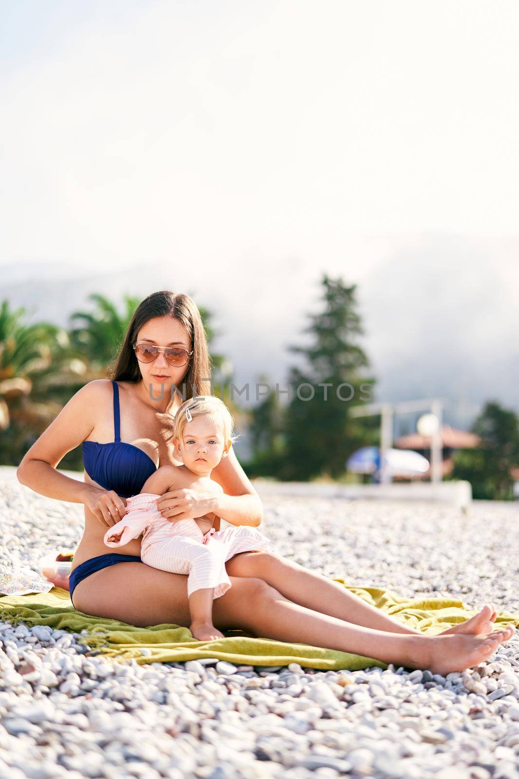 Mom in a bathing suit puts on a little girl a jumpsuit on the beach by Nadtochiy