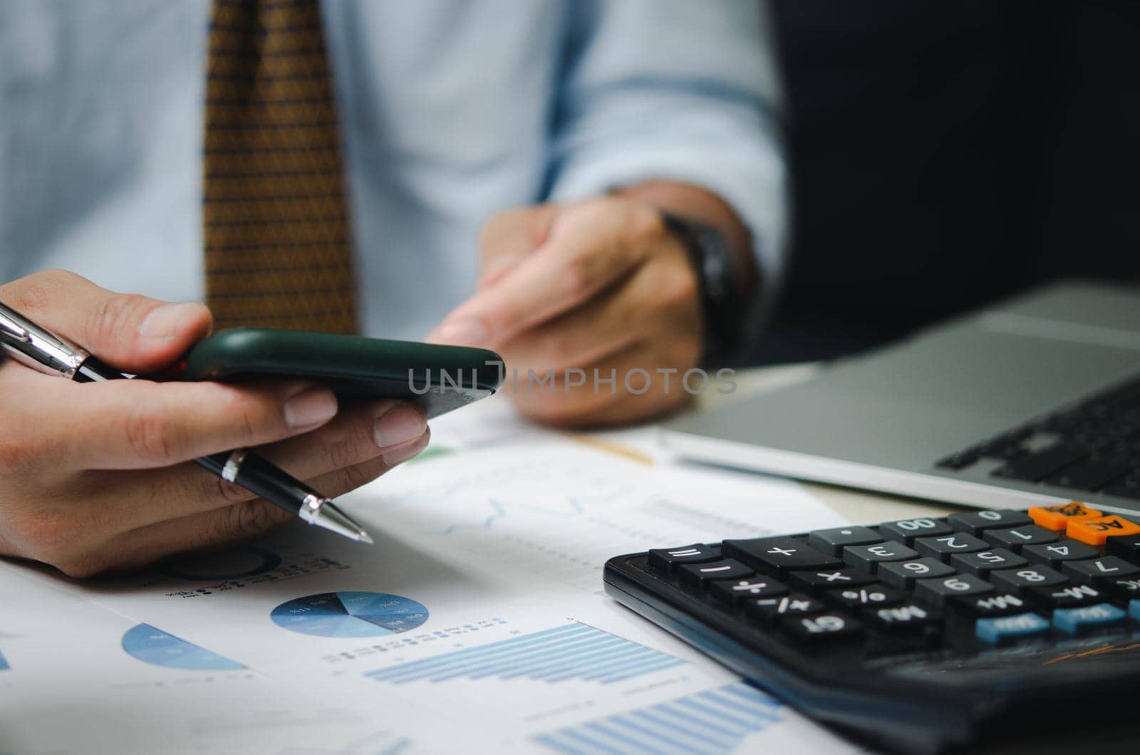 Businessman hand holding mobile phone and pen with graphs and charts business documents on desk. Calculator and computer for investment and economic analysis. Financial and tax business concept. by aoo3771