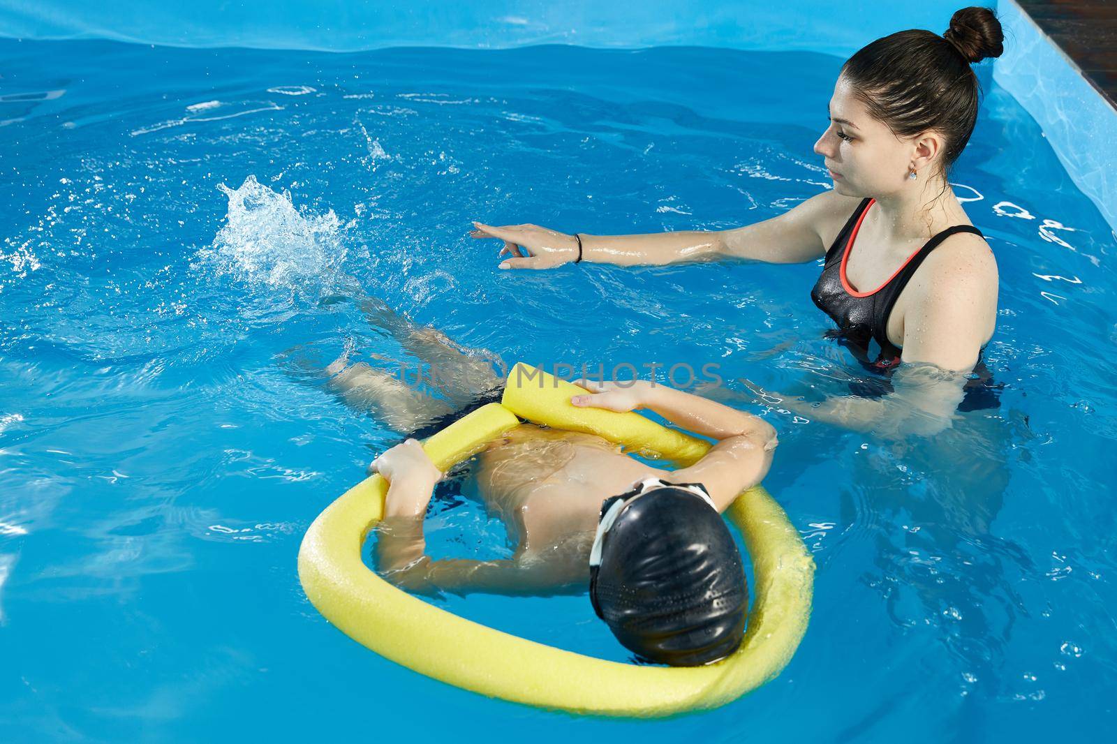Preschool boy learning to swim in pool with foam noodle with young trainer by Mariakray