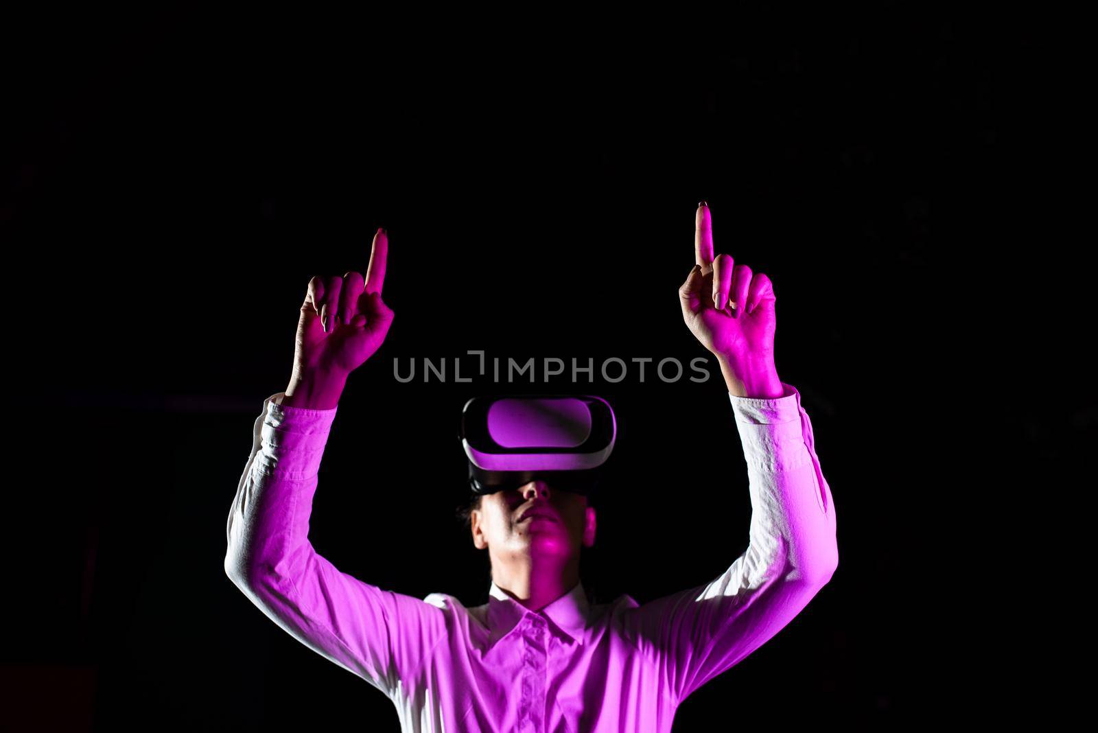 Female Manager Wearing Virtual Reality Goggles Raising Hands While Taking Professional Training. Light Falling On Woman Using Gadget And Presenting Futuristic Technology. by nialowwa