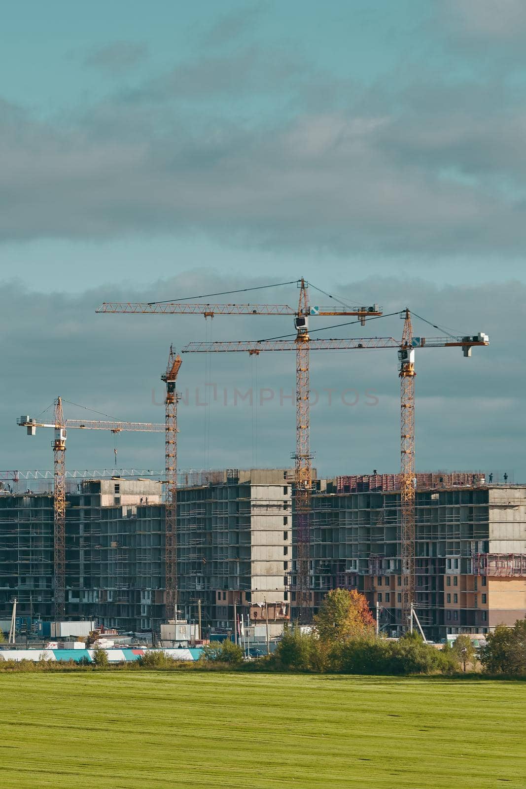 Construction site with industrial building cranes, multi-storey buildings of new city districts and large green field. Project of urban area. Blue sky with clouds.