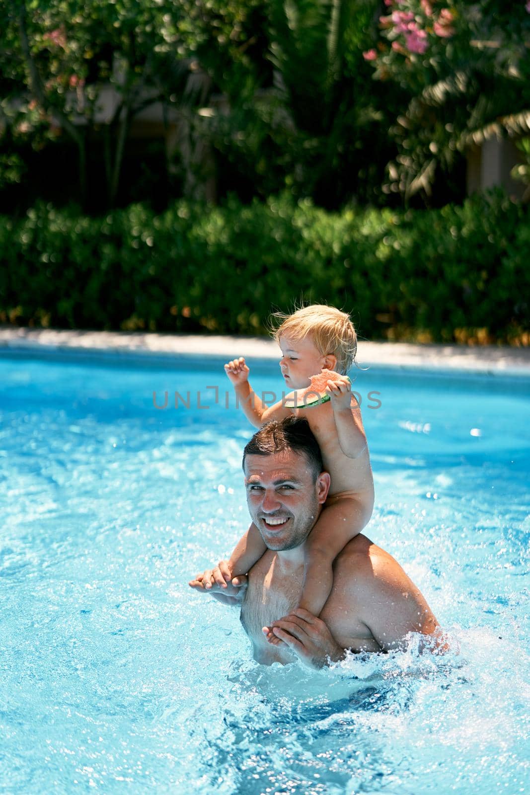 Little girl with a watermelon in her hand sits on the shoulders of her dad in the pool. High quality photo