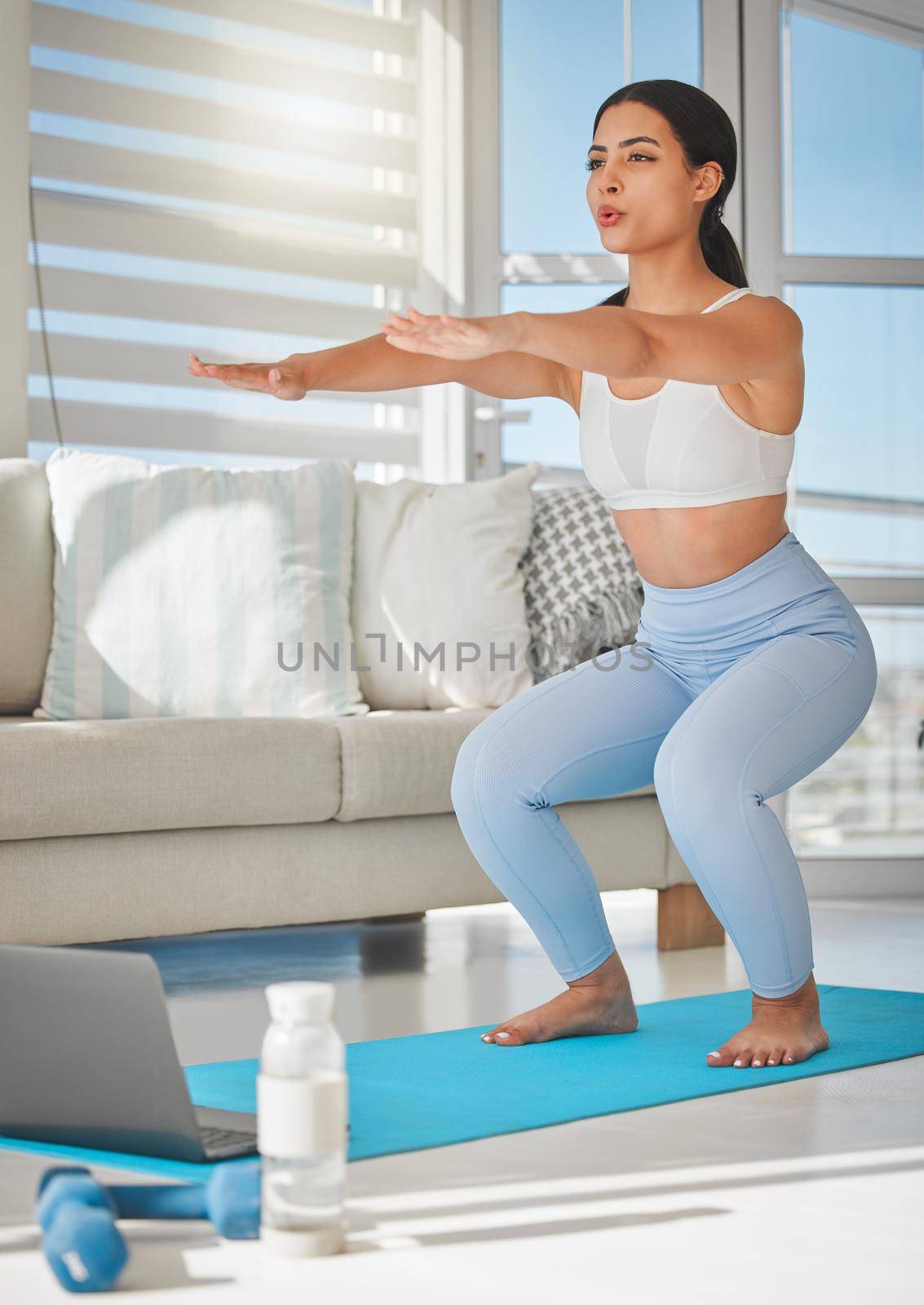 a sporty young woman using a laptop and doing squats while exercising at home.