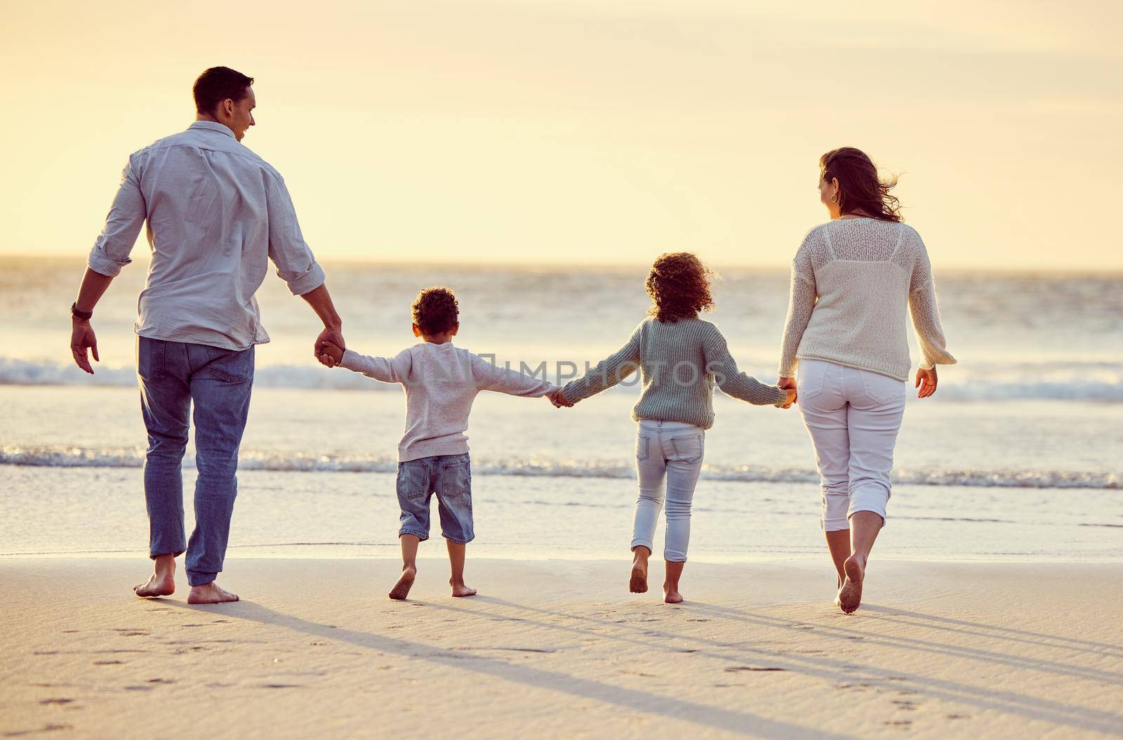 Rear view shot of a carefree family holding hands while walking on the beach at sunset. Mixed race parents and their two kids spending time together by the sea enjoying summer vacation.